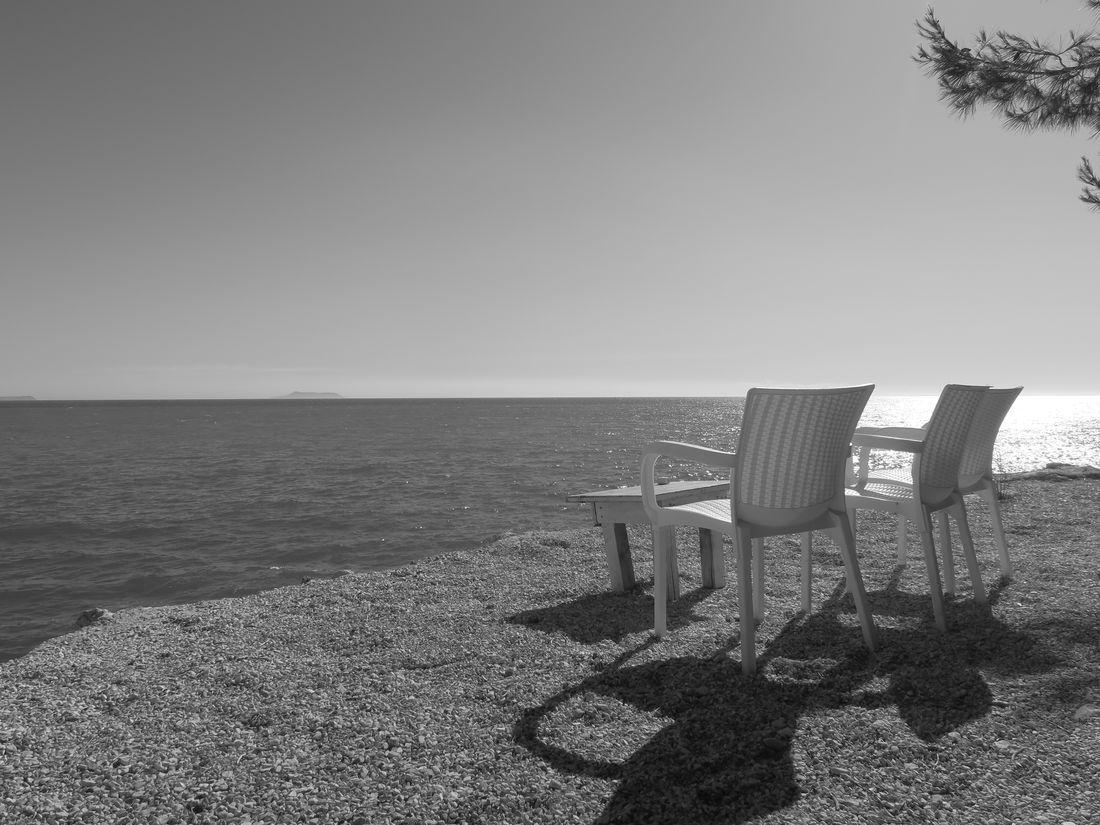 Zwei Stühle und ein Tisch an einem steinigen Ufer mit Blick aufs Meer, Schwarz-Weiß.
